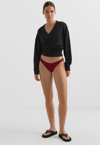 Černý top s dlouhými rukávy v zavinovacím stylu, žebrované burgundské tanga a černé žabky s tlustými podrážkami na jednoduchém bílém pozadí.