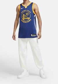 Královsky modrý basketbalový dres se žlutými akcenty, který nese nápis "Golden State Warriors" a logo. Doplněný bílými teplákovými kalhotami a teniskami.