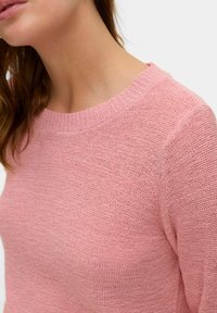Roza pletena jopica z okroglim izrezom, strukturirano tkanino in rebrastimi manšetami. Oblikovanje vsebuje ohlapno prileganje in subtilne šivane podrobnosti.