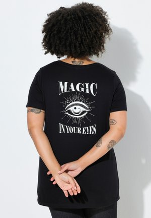 Persona con cabello rizado de pie mirando hacia una pared, lleva una camiseta negra con un gráfico de un ojo blanco y el texto "MAGIC IN YOUR EYES" en la espalda.