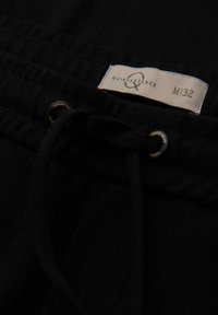 Gros plan sur un tissu noir avec une étiquette blanche portant la mention "Quintessence M32" et un cordon noir passant à travers des œillets en métal.