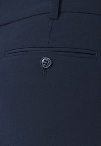 Granatowy materiał o gładkiej strukturze ma małą, skośną kieszeń zapinaną na okrągły czarny guzik z czterema otworami.