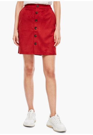 Jupe en denim rouge à coupe droite, avec cinq boutons à l'avant et une fente à l'arrière. Portée avec des baskets blanches pour un look décontracté.
