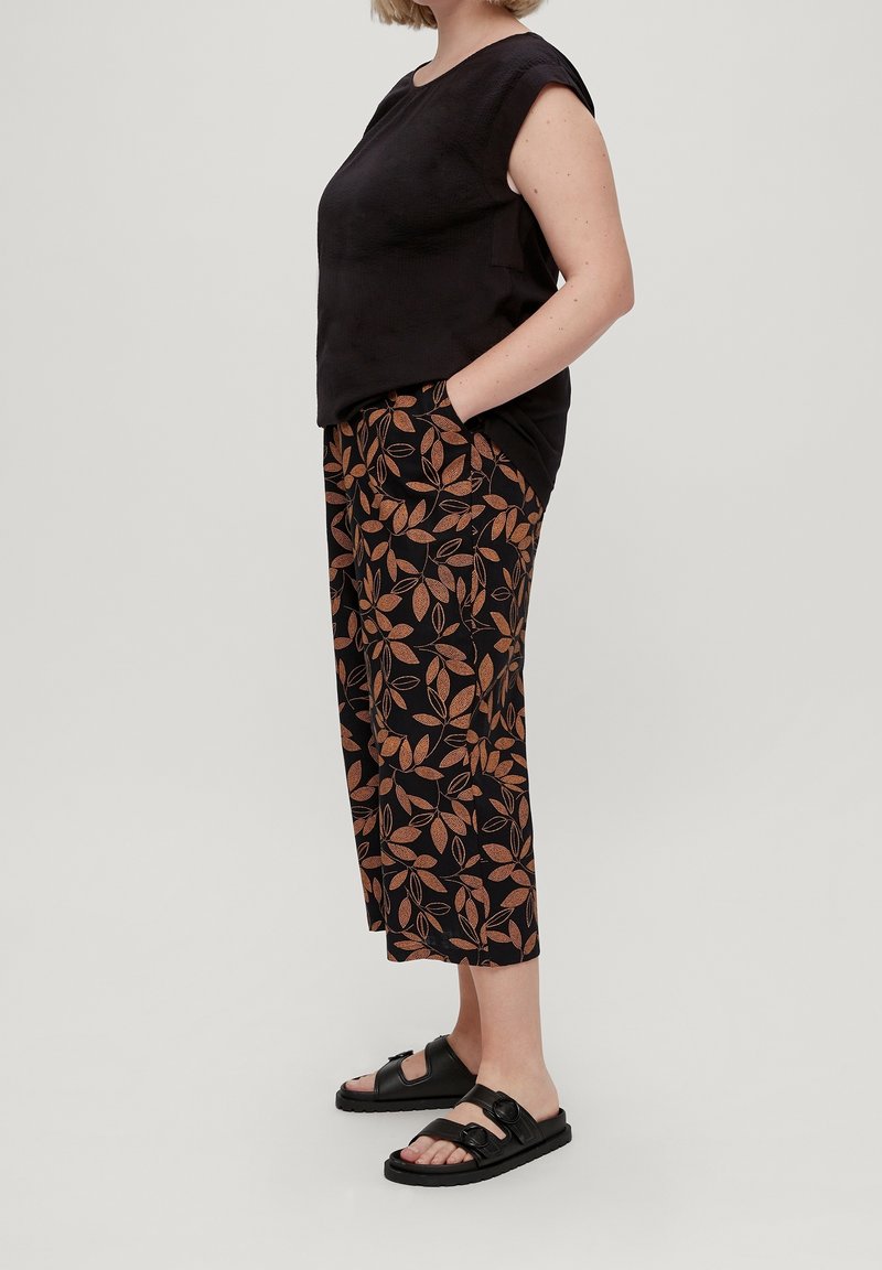 Femme portant un haut noir sans manches, un pantalon noir court avec un imprimé de feuilles marron, et des sandales noires ouvertes à l’avant, debout de profil.