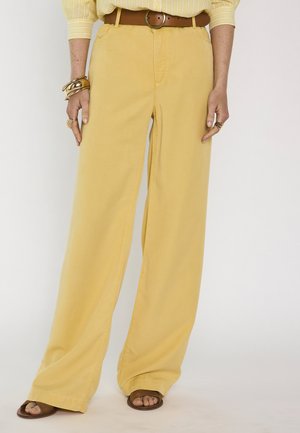 Femme portant un pantalon jaune à jambes larges, une ceinture marron, une chemise jaune rayée, des sandales marron, ainsi que des bracelets et des bagues en or aux mains.
