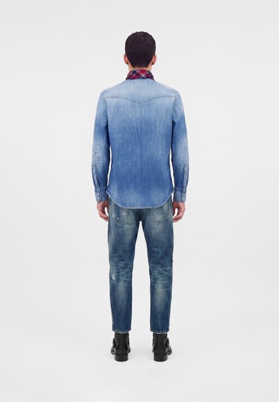 Man standing with back to viewer wearing faded blue denim shirt, distressed jeans, black boots, and red plaid collar visible.