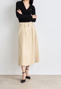 Cardigan noir à côtes avec un décolleté en V, associé à une jupe plissée beige avec une large ceinture et une boucle en argent. Chaussures à talons aiguilles noires avec bride à la cheville.