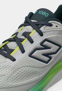 Γκρι αθλητικό παπούτσι με σκούρα κορδόνια, νέον πράσινες λεπτομέρειες, μεγάλο λογότυπο "N" στο πλάι και "1080" σε τιρκουάζ στο μαύρο γλώστρο.