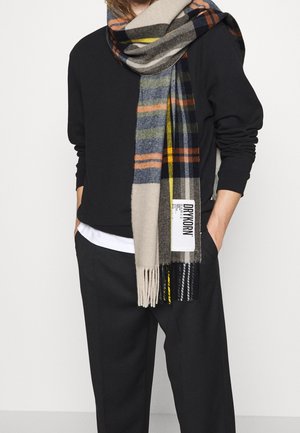 Personne portant un haut à manches longues noir et un pantalon, les mains dans les poches, avec une grande écharpe à carreaux beige, bleu, orange et noir enroulée autour du cou.