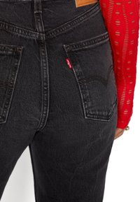 Los pantalones vaqueros de mezclilla negra presentan un diseño de cintura alta, dos bolsillos traseros con detalles de costura y una etiqueta roja en el bolsillo derecho.