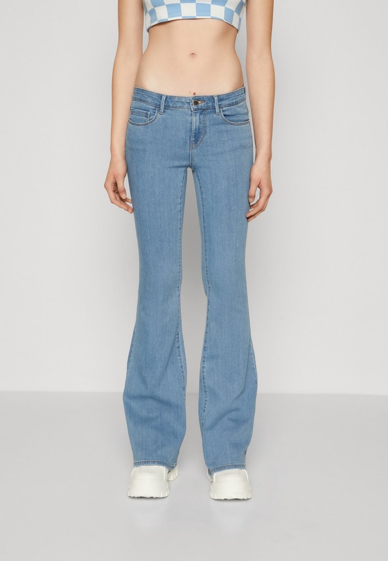 Femme portant un jean évasé bleu clair, des chaussures blanches épaisses et un crop top à carreaux bleu et blanc, debout devant un fond uni.