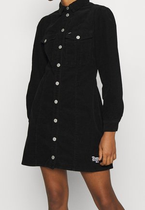 Czarna sukienka sztruksowa z zapięciem na guziki z przodu, kołnierzykiem, długimi bufkowymi rękawami, dwoma kieszeniami na piersiach oraz małym logo przy dolnym brzegu.