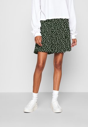 Vrouw die een wit top met lange mouwen draagt, een donkergroene rok met witte stippen, witte sokken en witte sneakers, staand op een grijze vloer.