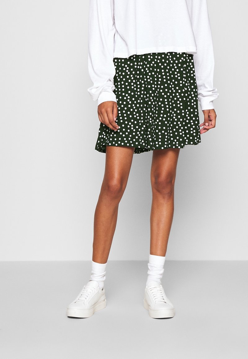 Femme portant un haut blanc à manches longues, une jupe vert foncé à pois blancs, des chaussettes blanches et des baskets blanches, debout sur un sol gris.