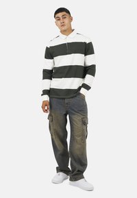 Polo a maniche lunghe con strisce verde scuro e bianche, abbinato a pantaloni cargo larghi e sbiaditi con multiple tasche e sneakers bianche.