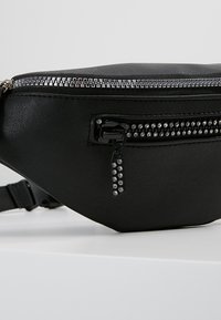 Mustakemmainen keinonahkainen vyölaukku, jossa on hopeanvärinen vetoketju, koristeelliset niittikoristeet ja säädettävä hihna. Kompakti, tyylikäs muotoilu.