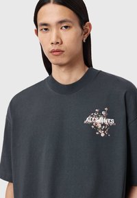 Σκούρο γκρι κοντομάνικη μπλούζα από απαλό ύφασμα, με φυτικά γραφικά και εκτύπωση κειμένου στο μπροστινό μέρος. Στρογγυλό λαιμόκοψη.