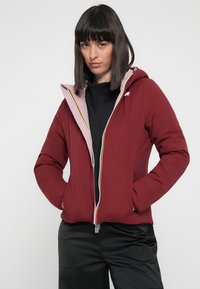 Maroon puffer jacket with a light pink hood lining, full zipper, side pockets, elastic cuffs, and a logo detail on the chest.