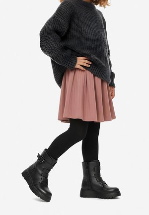 Stivali combat neri con fibbie decorative, gonna plissettata rosa e maglione oversize testurizzato grigio scuro. Modella con collant neri.