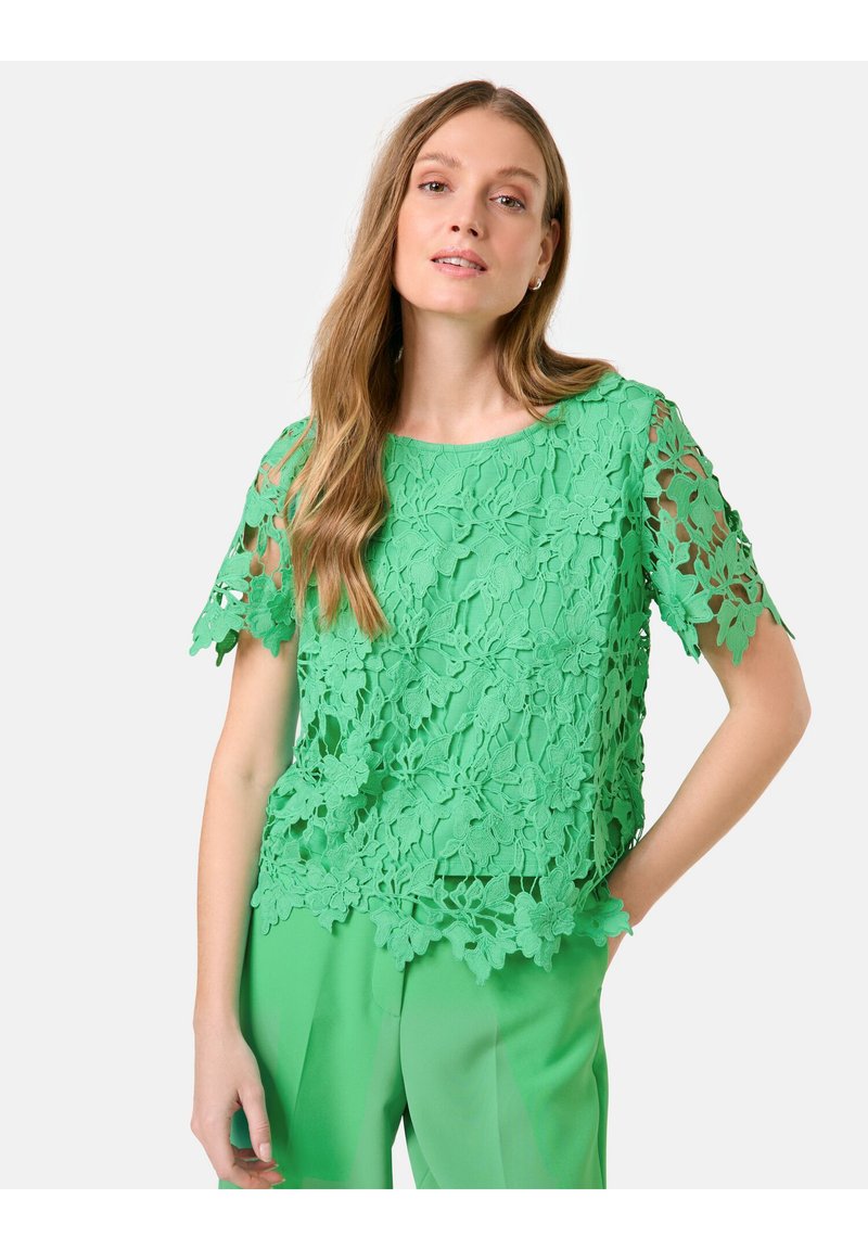 Grüner Spitzenoberteil mit floralem Muster, kurzen Ärmeln und rundem Ausschnitt, kombiniert mit passenden grünen Hosen. Strukturiertes Design.