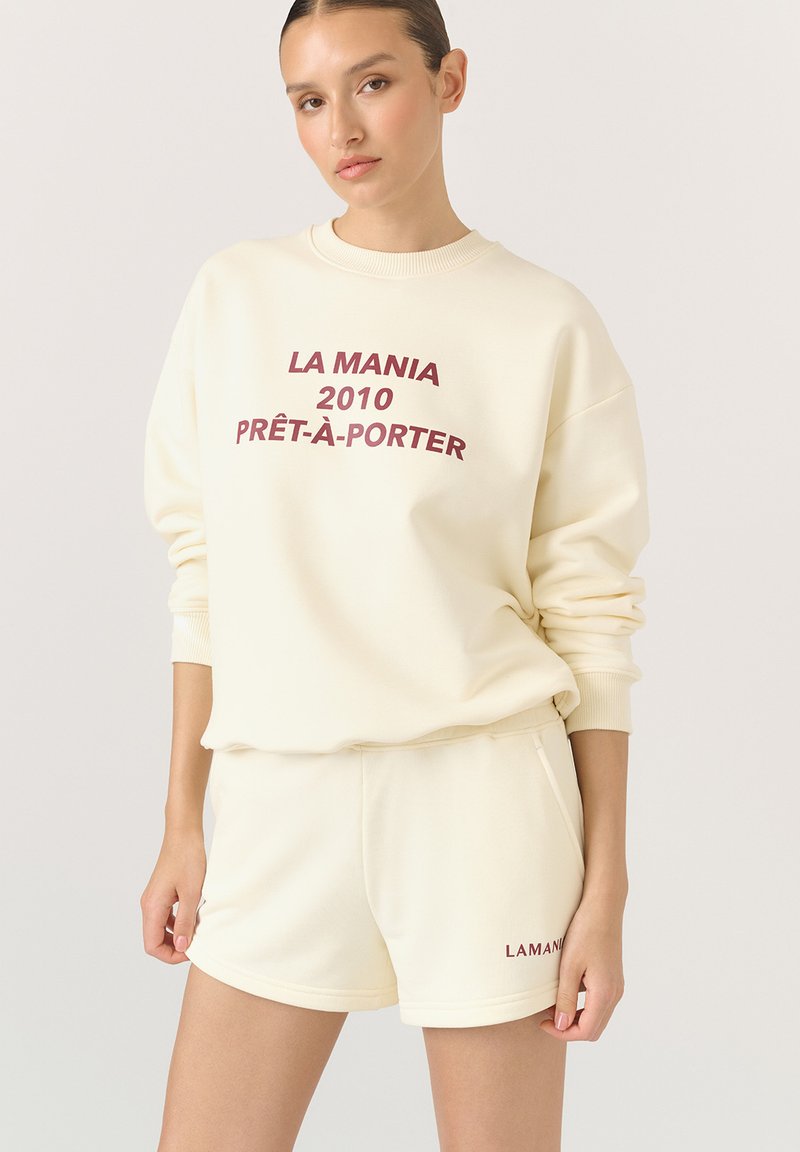 Zestaw bluzki i szortów w kolorze kremowym. Bluzka ma bordowy napis "LA MANIA 2010 PRÊT-À-PORTER". Gładki materiał, luźny krój, ściągacze na rękawach.