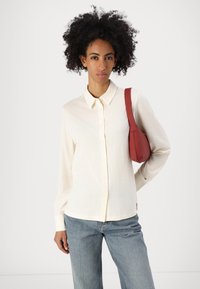 Kremfarget langermet skjorte med knappestolpe, lett tekstur, kombinert med lyseblå jeans og en rød veske. Enkel, strukturert design.