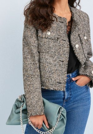 Femme portant une veste en tweed texturé, un top en dentelle noire, un jean bleu, tenant un sac à main vert doux avec une bandoulière en chaîne argentée.