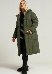 Olivgrön, lång pufferjacka med quiltad design, huva och sidofickor; matchad med svarta byxor och ankelstövlar.