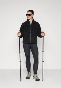 Žena oblečená v černé fleecové bundě, černých legínách, šedých turistických botách a tmavých slunečních brýlích, držící pár černých turistických holí.