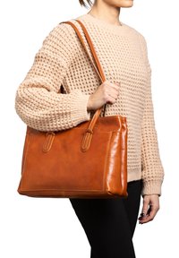 Tote bag van tan leer met dubbele handvaten, rechthoekige vorm, contrasterende stiksels en een soepele textuur, vastgehouden door een persoon in een gebreide trui.