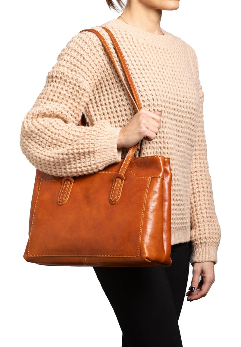 Tote bag van tan leer met dubbele handvaten, rechthoekige vorm, contrasterende stiksels en een soepele textuur, vastgehouden door een persoon in een gebreide trui.