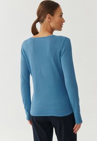 Blaues Langarm-Oberteil aus geripptem Strick mit runder Halslinie und leicht geschwungenem Saum, das weiche Textur und eine figurbetonte Silhouette zeigt.
