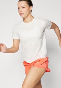 Camiseta atlética blanca de manga corta, con un pequeño logo. Combinada con unos pantalones cortos de color coral brillante diseñados para el movimiento, mostrando una textura suave.