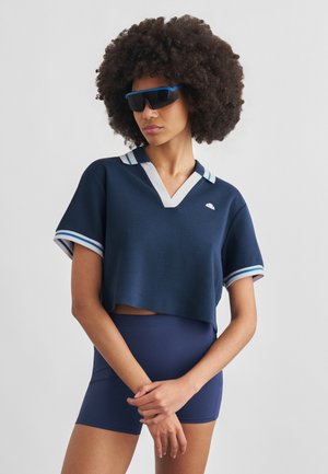 Nagy göndör hajjal rendelkező nő fekete sport napszemüveget visel, sötétkék, fehér szegélyű rövid felsőt és sötétkék, testhezálló rövidnadrágot visel.