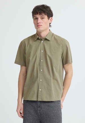 Jonge man draagt een lichtgroen overhemd met korte mouwen en donkere grijze spijkerbroek, staand tegen een effen lichte achtergrond.