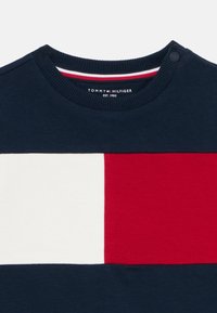 Námornícky modrý sveter s okrúhlym výstrihom s veľkými bielymi a červenými obdlžníkovými farebnými blokmi a s nálepkou Tommy Hilfiger na golieri.