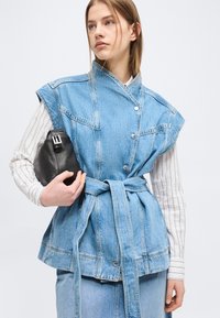 Νεαρή γυναίκα με ίσια ανοιχτό καστανή μαλλιά φοράει αμάνικο τζιν γιλέκο με ζώνη πάνω από μακρυμάνικο ριγέ πουκάμισο, κρατώντας μαύρο τσαντάκι χειρός.