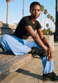 Noor inimene musta särgi ja siniste pükstega istub välitingimustes puupingil jalad ristis, taustal palmid.