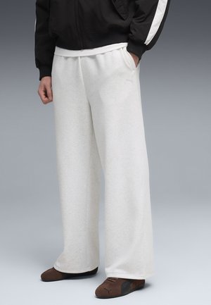 Persoon draagt losse witte joggingbroek, zwart-witte jas en bruine Puma-schoenen, staat met één hand in de zak.