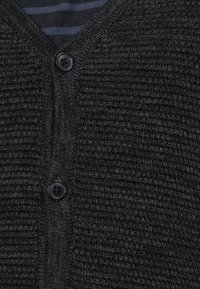 Svart stickad cardigan med V-hals, två svarta knappar och ett texturerat mönster. Bakgrundsfabriken har marinblå ränder.