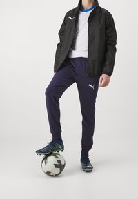 Černá větrovka, bílá tričko, námořnické tepláky a modré kopačky na fotbal. Stojí na černobílém fotbalovém míči.