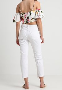 Kvinna som bär vita högmidjade jeans med fransade nederkanter och en blommig av-axeln crop top med volanger, som står mot en enfärgad bakgrund.