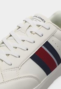 Λευκό αθλητικό παπούτσι με λευκά κορδόνια, λωρίδα από ύφασμα μπλε σκούρου και κόκκινου χρώματος στο πλάι, και το λογότυπο Jack & Jones στη γλώσσα.