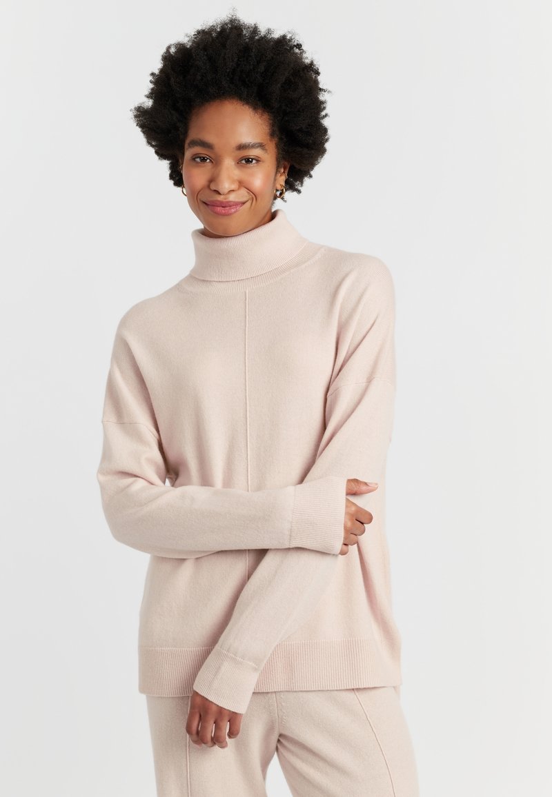 Pull en col roulé rose clair en tissu tricoté doux, avec des épaules tombantes, des manches longues et une coupe décontractée. Poignets et ourlet côtelés.