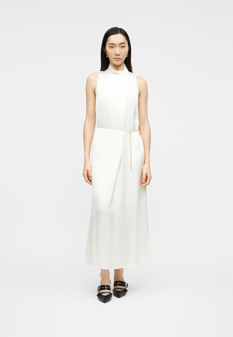 Γυναίκα που στέκεται φορώντας ένα λευκό αμάνικο φόρεμα με ψηλό γιακά, λεπτή ζώνη και μαύρα μυτερά παπούτσια με λουριά και τρύπες για κορδόνια.