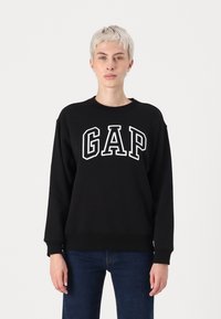 Juodas medvilnės džemperis su apvaliu kaklu, kuris turi baltą "GAP" logotipą per krūtinę. Ilgos rankovės ir gumbuoti rankogaliai.