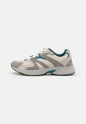 Zapatilla deportiva beige y gris con cordones blancos y detalles en verde azulado, suela acolchada, vista desde el lado exterior sobre un fondo blanco.
