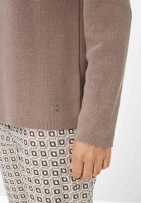 Pull en tricot beige avec un ourlet côtelé, associé à un pantalon à motifs présentant des designs carrés dans des couleurs atténuées. Accent logo sur le pull.