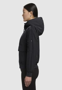 Veste à capuche noire avec une finition brillante, des poignets élastiques et une poche avant ; présente des cordons au niveau de la capuche et un design court.