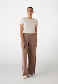 T-shirt in cotone beige chiaro con un logo all'interno di un design triangolare, abbinato a pantaloni taupe a vita alta e a gamba larga.
